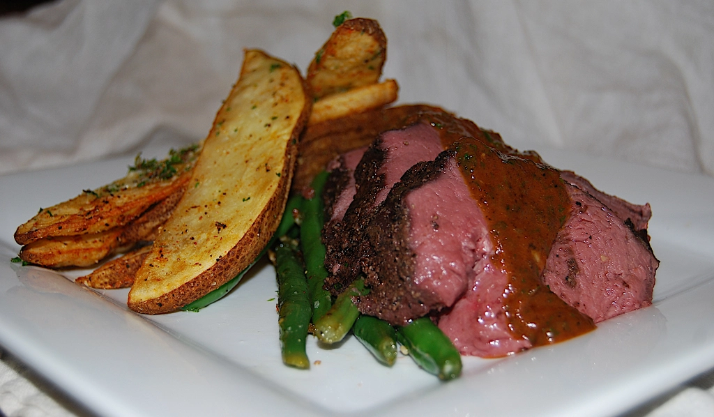 venison roast slices topped with red wine sauce with potato wedges and asparagus on a white plate.