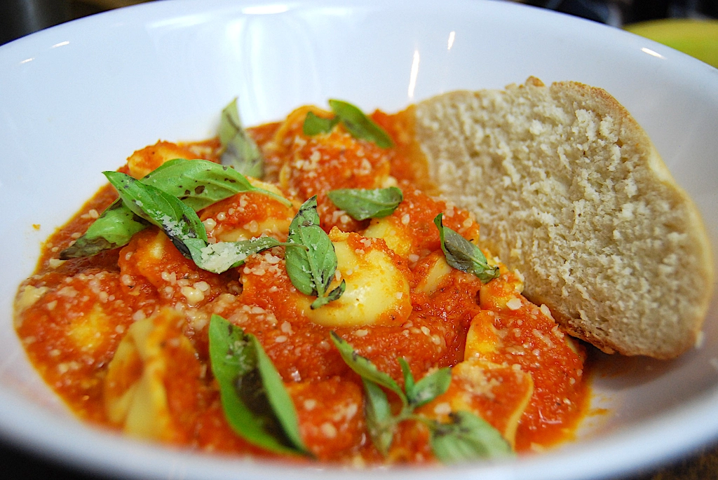 Tomato soup with carrot, leek, and tortellini soup in a white bowl with a piece of bread