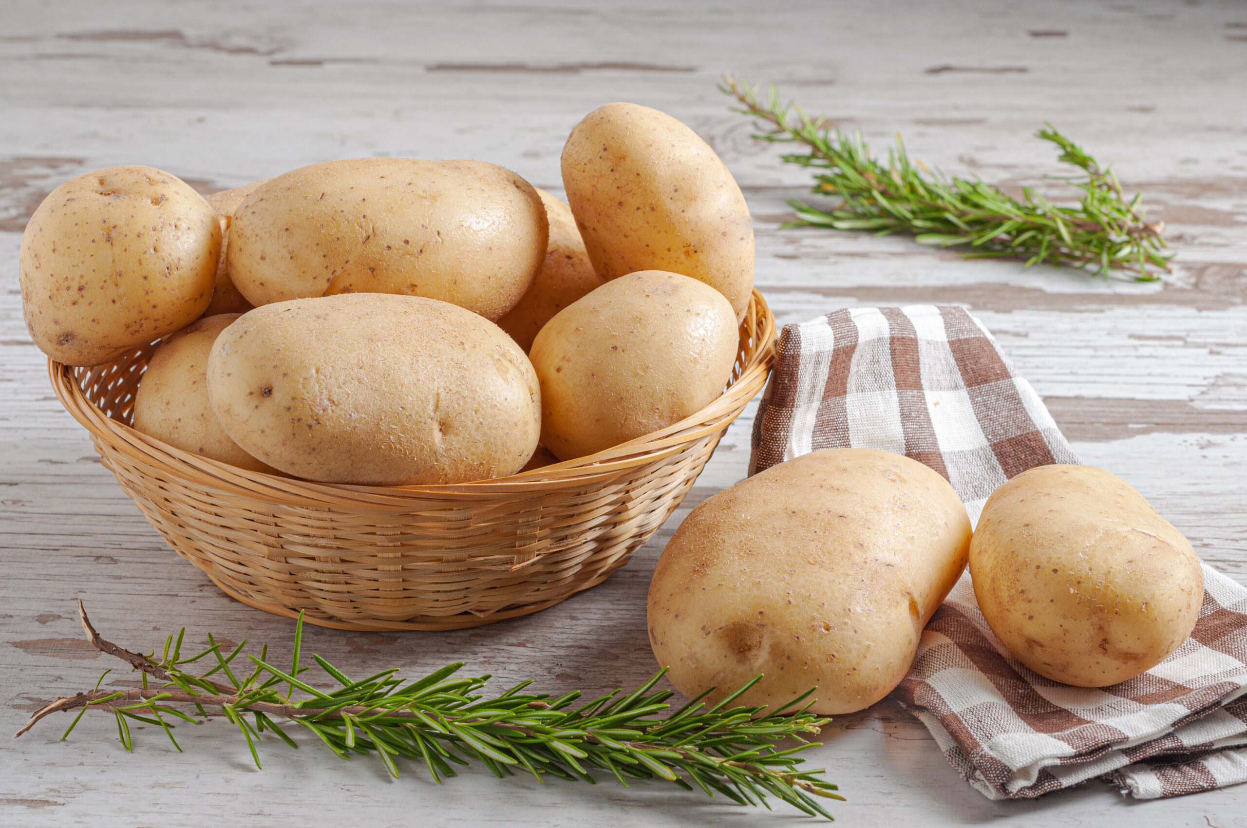 Fresh raw potatoes in a woven wicker basket with natural rosemary leaves on a wooden rustic table surface