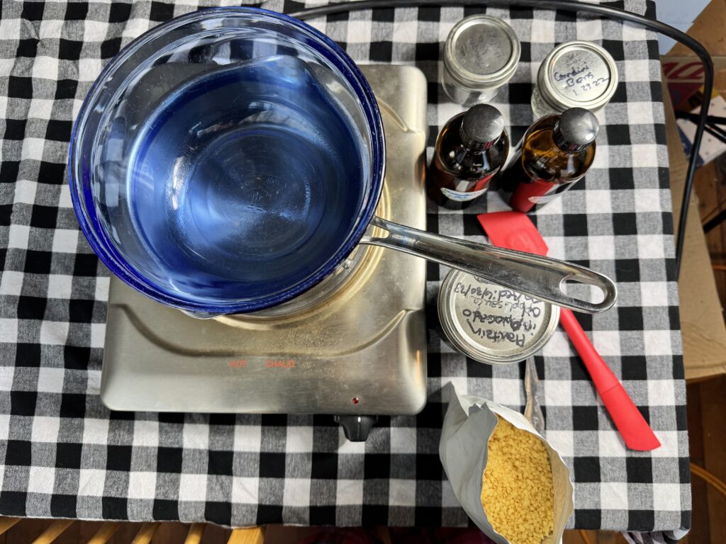 overview shot of DIY healing salve ingredients and tools: bain-marie, pelletized beeswax, red spatula, pint jar of plantain infused oil, brown bottles of calendula and comfrey infused oils, 2 4 oz jelly jars all laid out on a black checkered table cloth