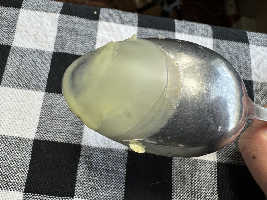 metal spoon with a coating of the DIY healing salve on the back of it being held over a black and white checkered table cloth
