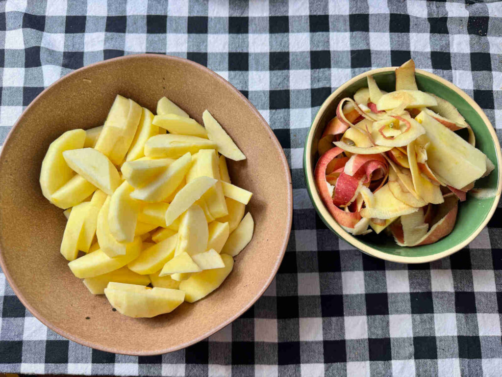 a brown bowl filled with apple slices and a green and yellow bowl filled with apple peels and cores, both side by side on a black and white checkered cloth