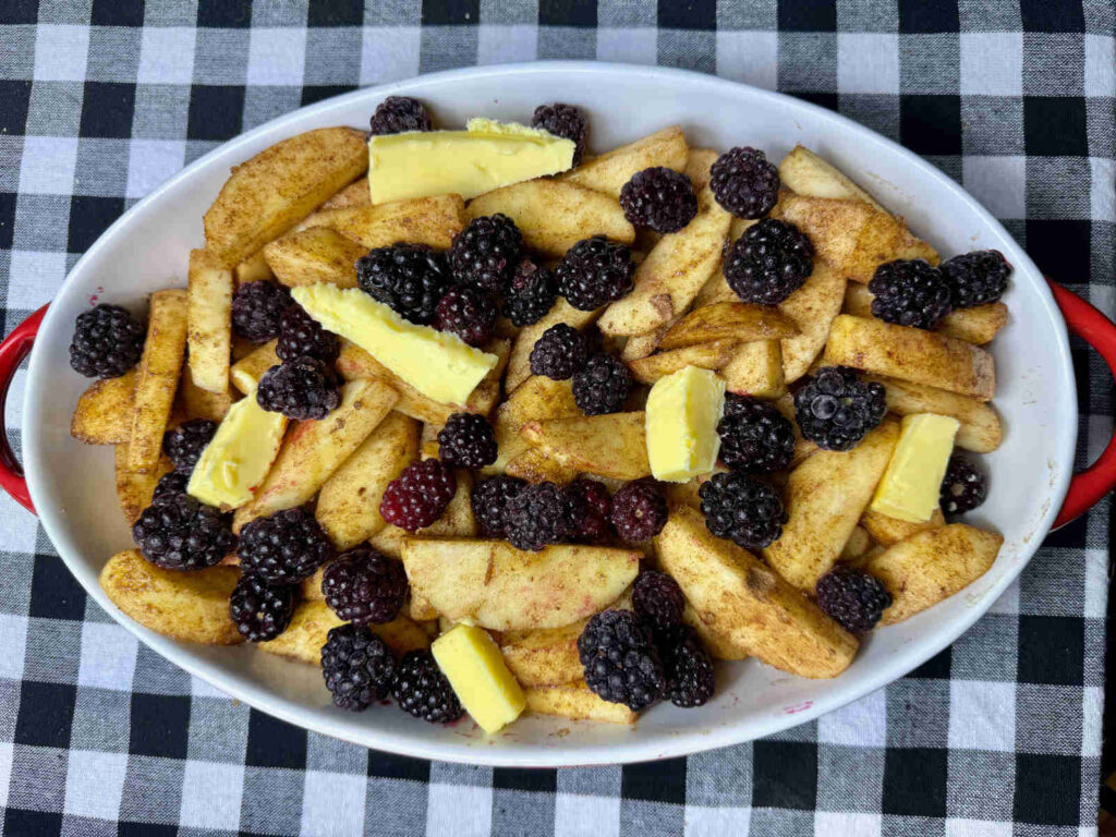 cinnamon covered apple slices, blackberries, and butter chunks in a white and red casserole dish on a black and white checkered cloth