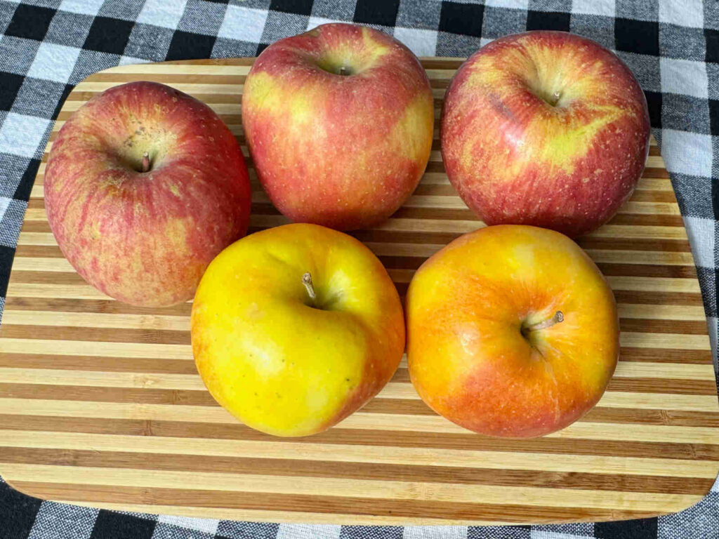 Five red and yellow apples on a light and dark brown striped cutting board