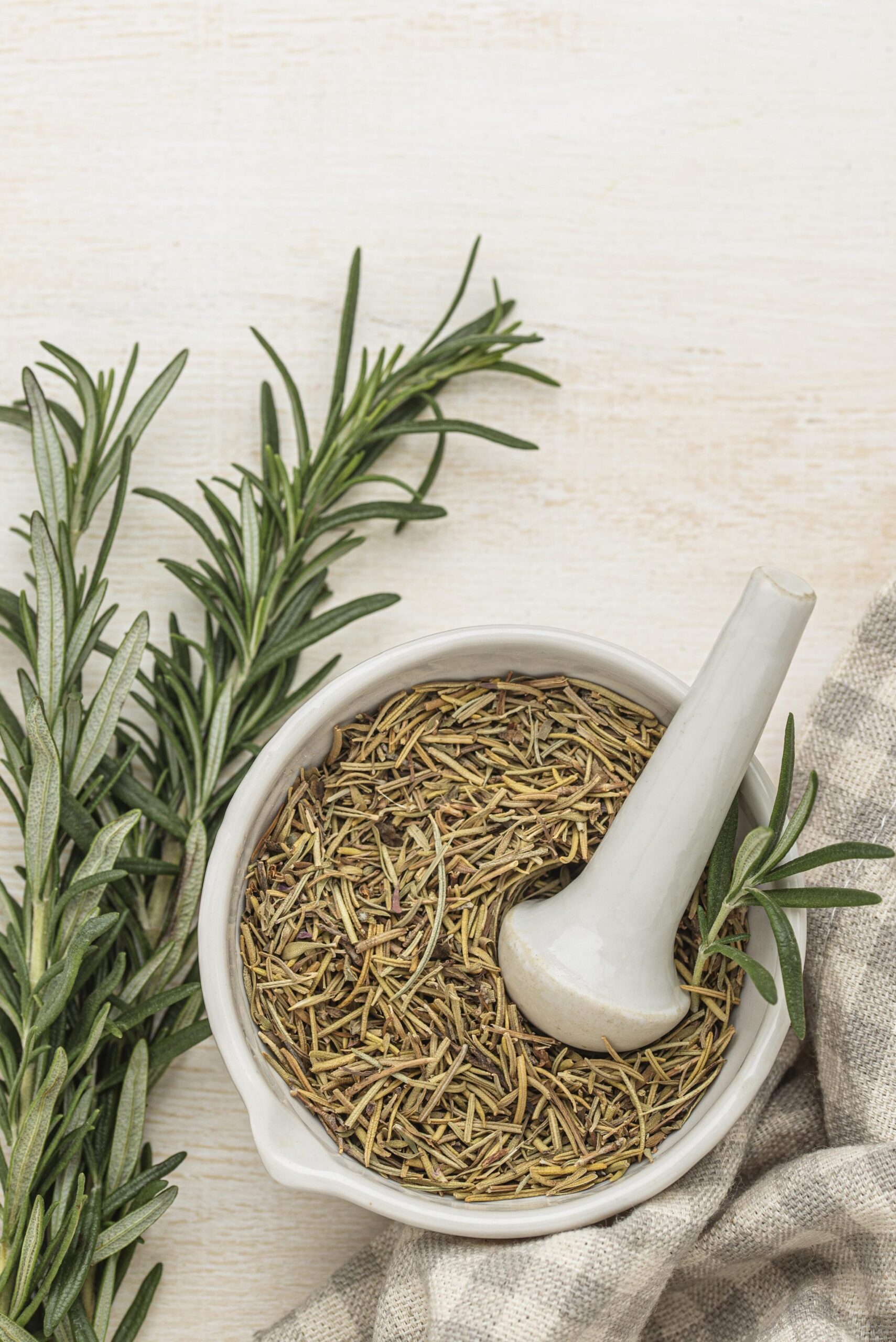 top view of dried rosemary in a mortar and pestle with a couple of fresh rosemary sprigs to the left and a bunched up gray and white gingham towel on the right.