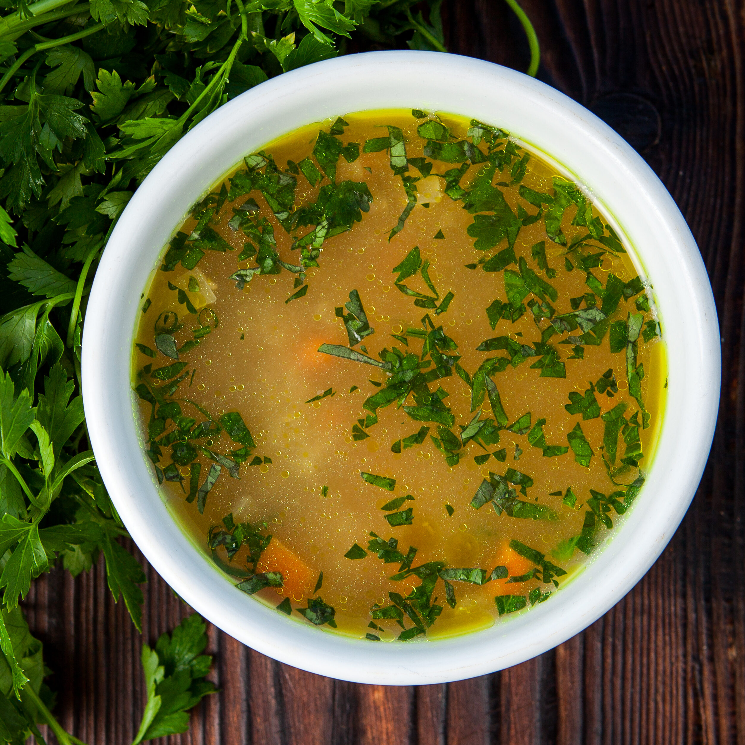 Top view soup with made with chicken bone broth, vegetables, and parsley in a white plate on a dark wooden background