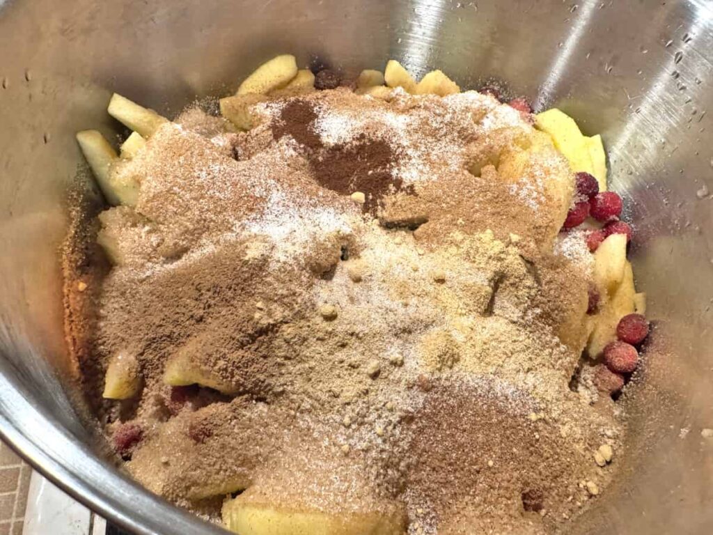 Sugar and spices on top of apple slices and cranberries in a stainless steel stock pot