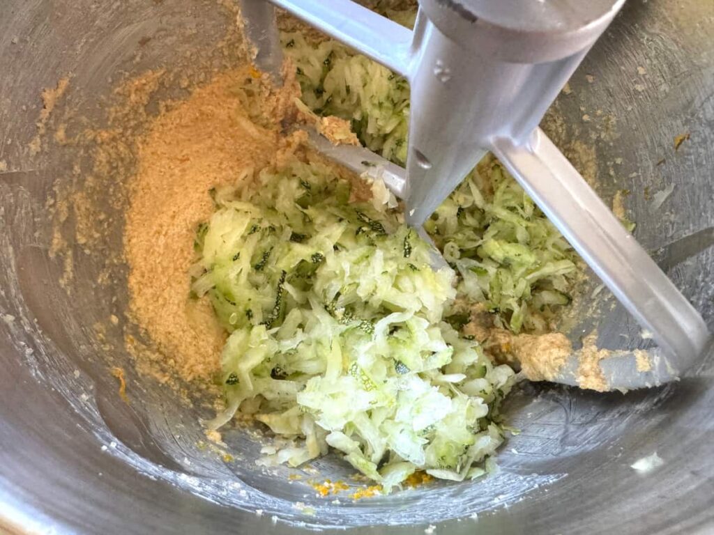 wet ingredients mixed together with shredded zucchini added in a stainless steel stand mixer bowl with a paddle attachment
