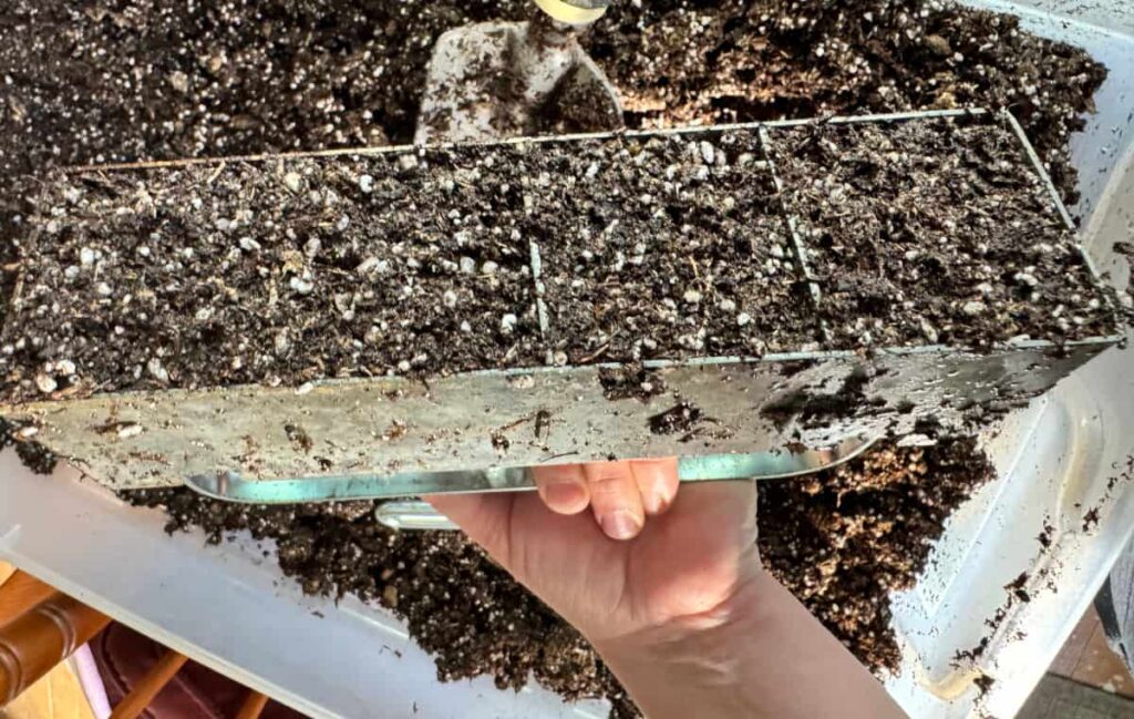 hand holding a soil blocker upside down over a white tub of soil with soil packed inside it to form blocks