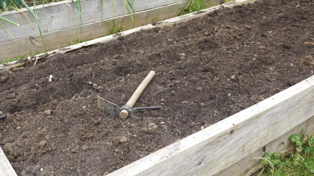 wooden raised garden bed filled with soil with a small hoe and cultivator garden tool sitting on top